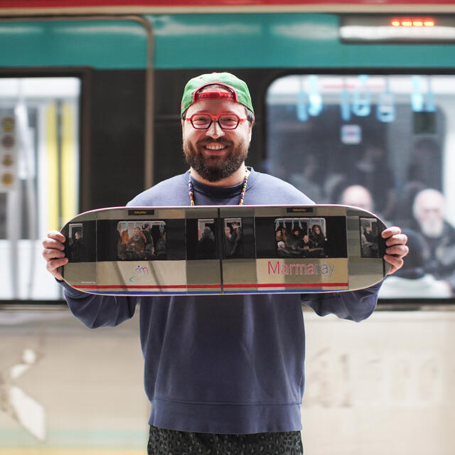 A picture of Berk Armağan holding their skateboard in front of a train station 
